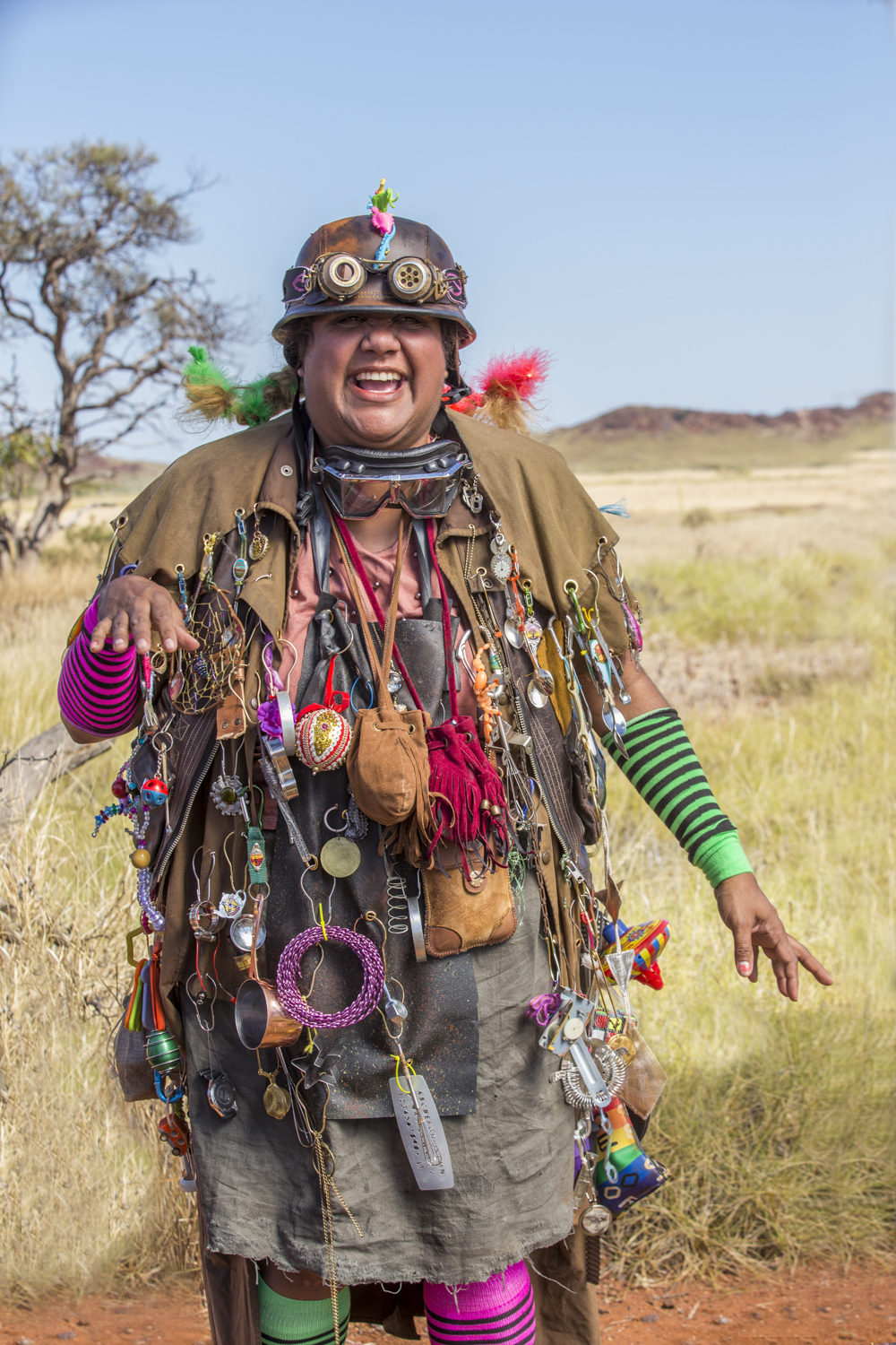 A woman wears steampunk inspired helmet, goggles and jacket adorned with hanging objects, stands and laughs in desert landscape.