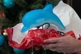Hands hold gift wrap containing a lamp in the shape of a dolphin. Christmas tree in background.