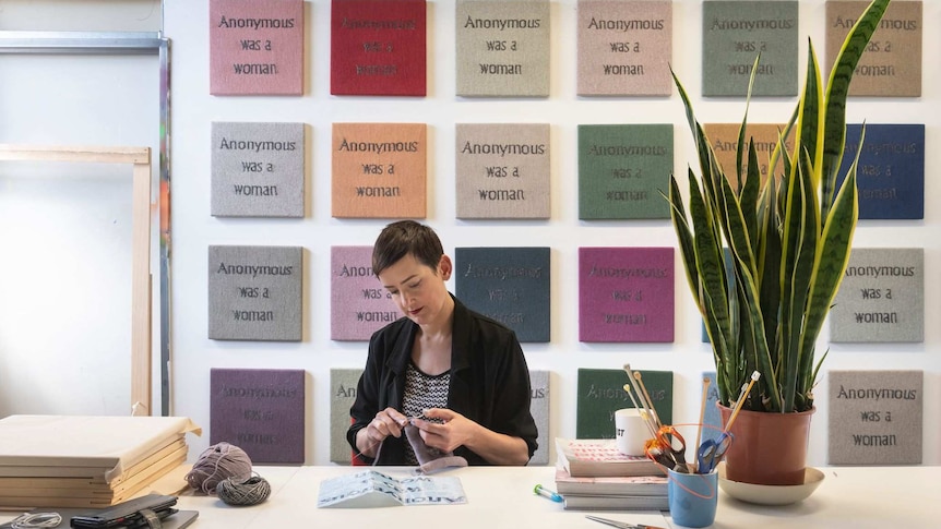 The artist Kate Just knitting with panels in the background that read "anonymous was a woman"