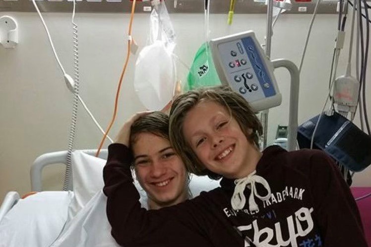 Two teenage boys smile as one lies in a hospital bed