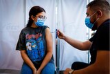 A young woman in a rock band t-shirt, wire framed glasses and a face mask gets a needle in her shoulder
