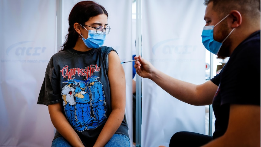 A young woman in a rock band t-shirt, wire-framed glasses and a face mask gets a needle in her shoulder