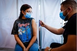 A young woman in a rock band t-shirt, wire framed glasses and a face mask gets a needle in her shoulder