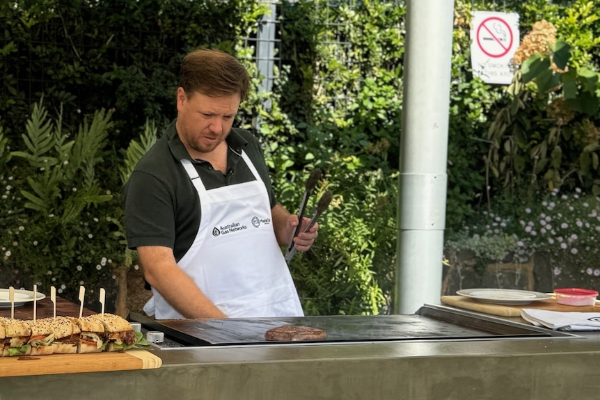 A man wearing a black polo shirt and white apron turns off a barbecue after cooking a steak.
