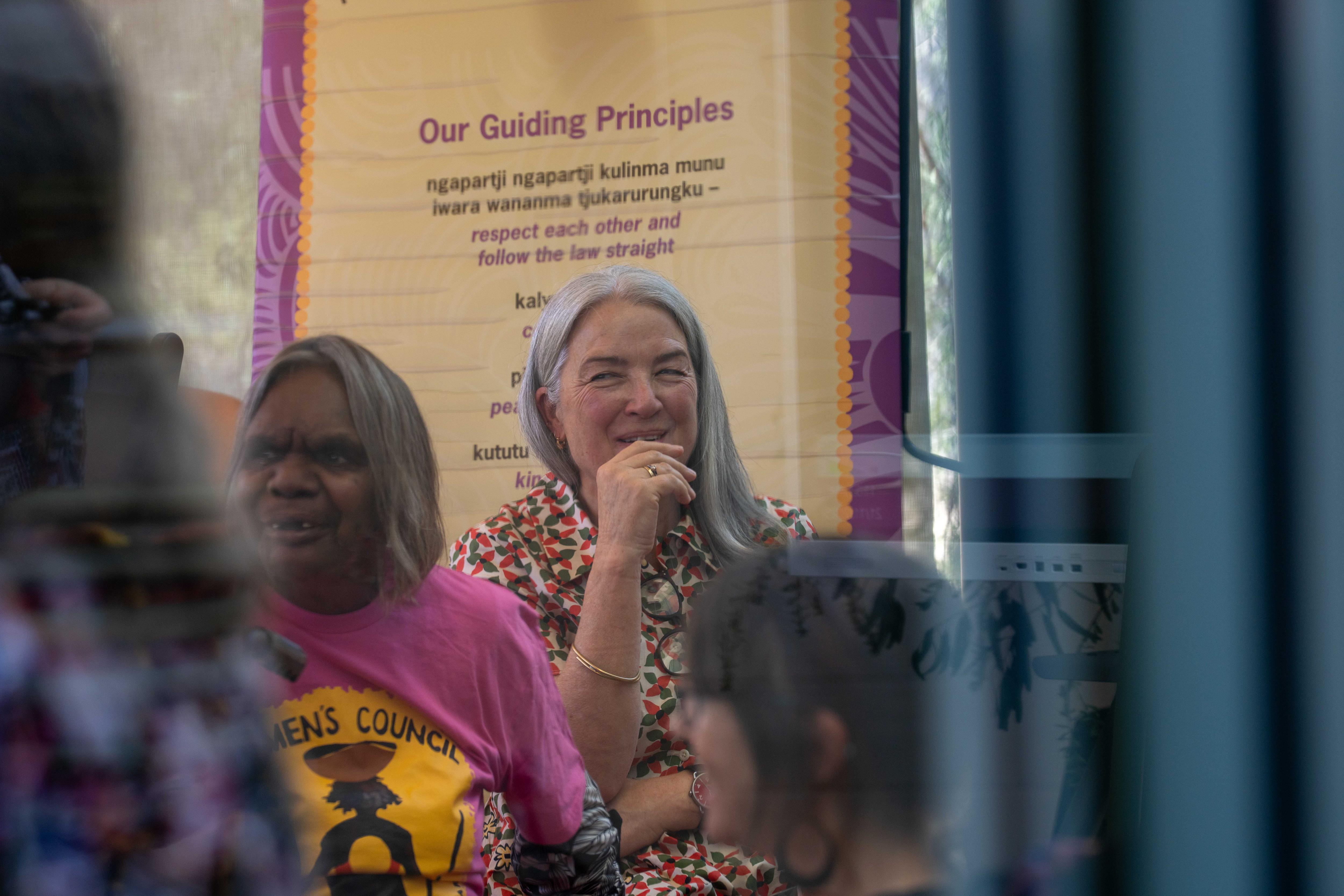Una señora de pelo gris es fotografiada a través de una ventana de cristal. Se lleva la mano a la barbilla y sonríe sin mirar a la cámara.