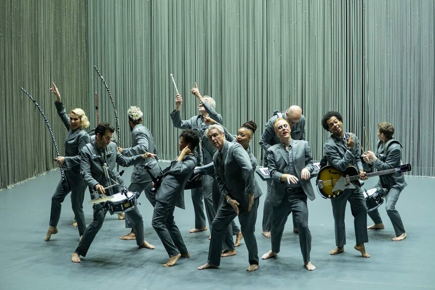 12 barefoot performers in grey suits dance on stage with metallic curtain perimeter, some with instruments and some without.