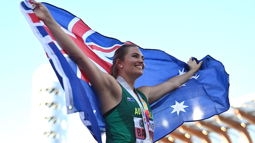 Kelsey-Lee Barber smiles while holding up the Australian flag and wearing a gold medal around her neck