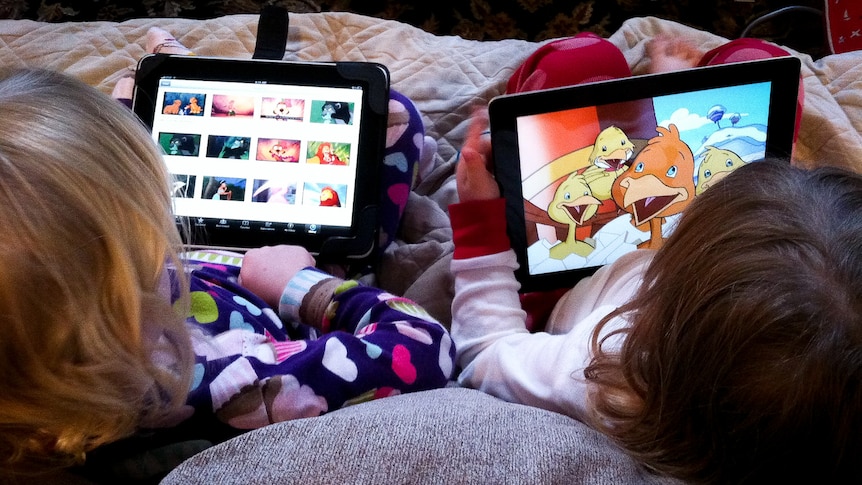 Two small children are sitting on a couch in colourful pajamas, watching cartoons on their tablets.
