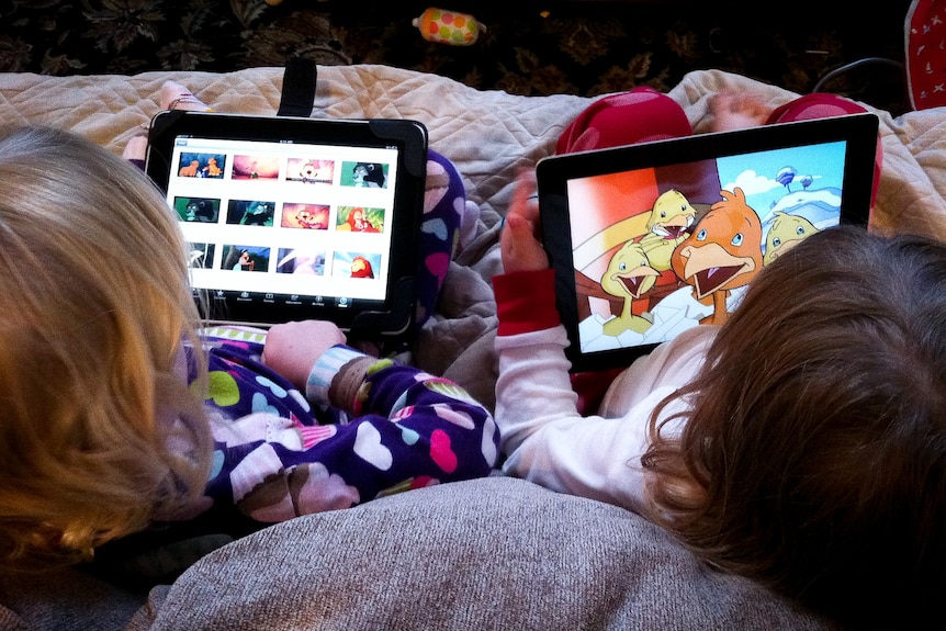 Two small children are sitting on a couch in colourful pajamas, watching cartoons on their tablets.
