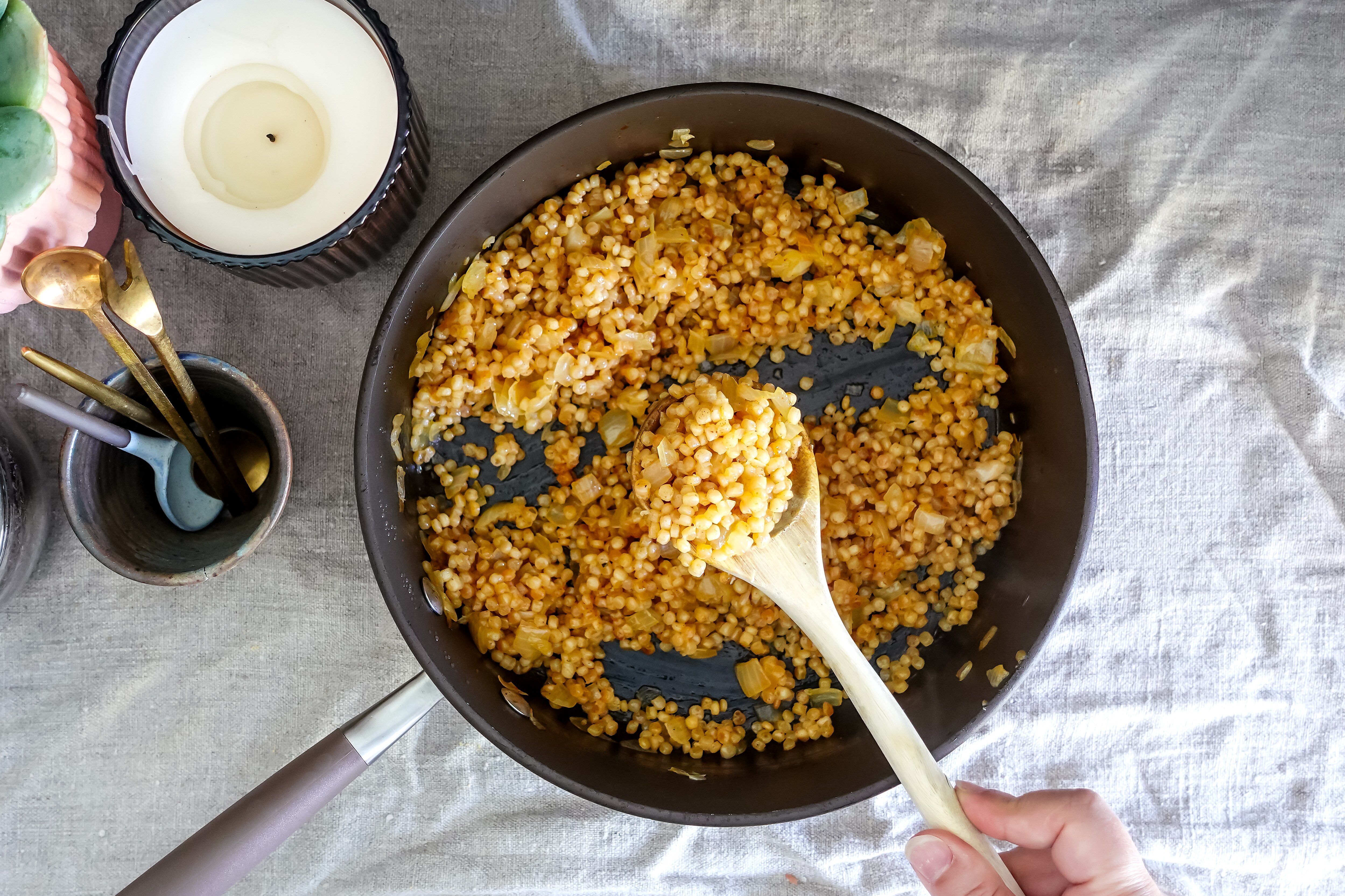 A frying pan with just-cooked pearly couscous and onion. A wooden spoon shows the cooked pearl couscous.