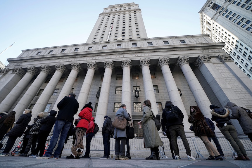 A line of people in winter coats stand outside a court building in New York City. The sky above them is clear