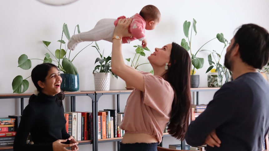 Woman holds up a baby in loving way while little sister laughs and dad watches on