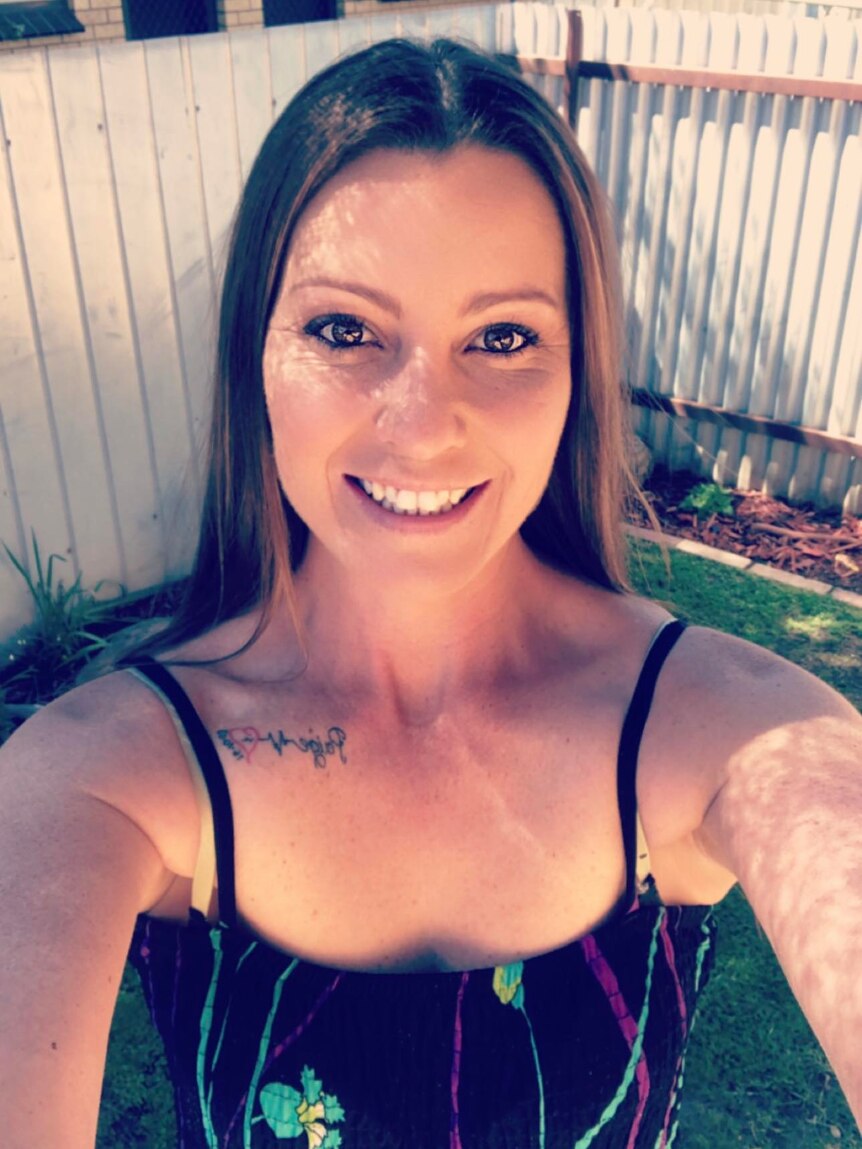 An image of a woman in her 30s taking a selfie while smiling, dressed in a singlet top with a neck tattoo.