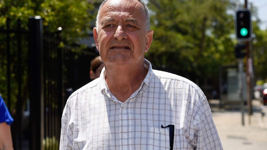 An older, bald man wearing a light-coloured shirt walks down a sunny street.