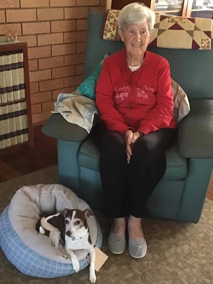 An elderly woman sits on an armchair with a small brown and white dog curled up at her feet.