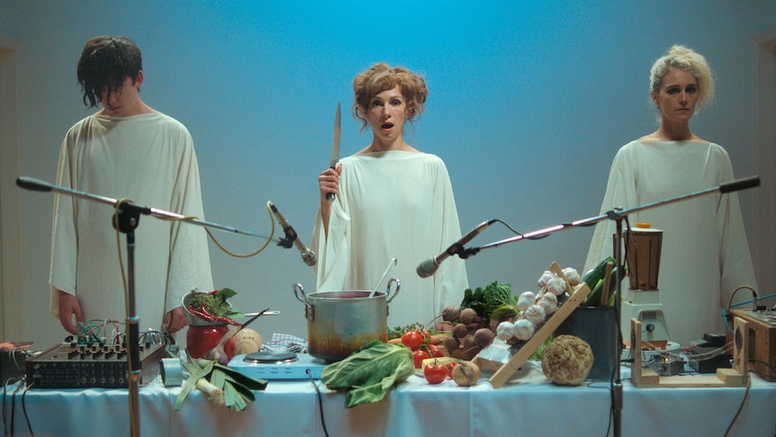 Three figures in white robes stand in front of a table laden with food. The woman in the middle holds up a knife