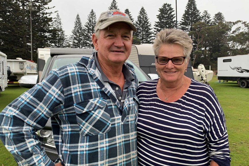 An older husband and wife couple stand in what looks like a caravan park in  coastal area.