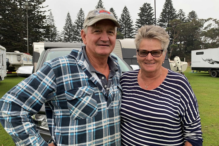 An older husband and wife couple stand in what looks like a caravan park in  coastal area.