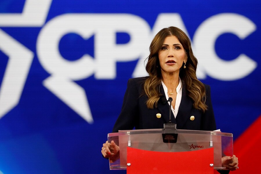 A middle-aged white woman with long dark hair and blonde highlights speaks behind a lectern in front of a bright background.