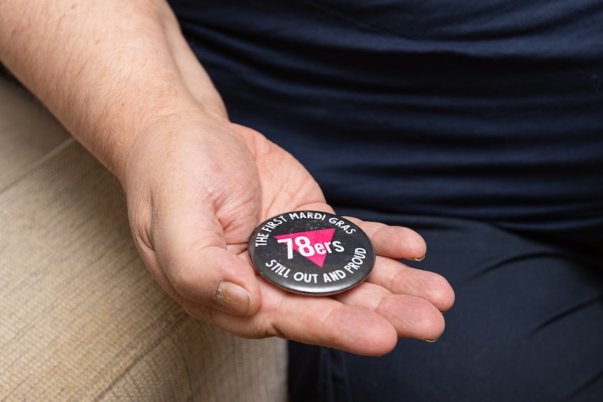 Peter's hand is seen holding a black and pink badge reading "78ers, the first Mardi Gras, still out and proud."