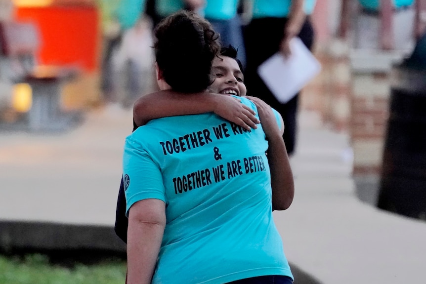  A teachers hugs a student. The back of her shirt reads "together we rise and together we are better". 