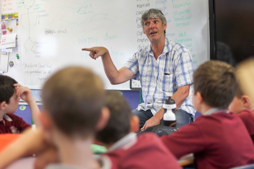 A teacher sits at the front of a classroom of kids, in front of a whiteboard, pointing.