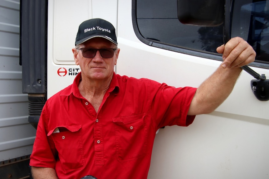 A man in a red shirt, cap and sunglasses leans against a truck. 