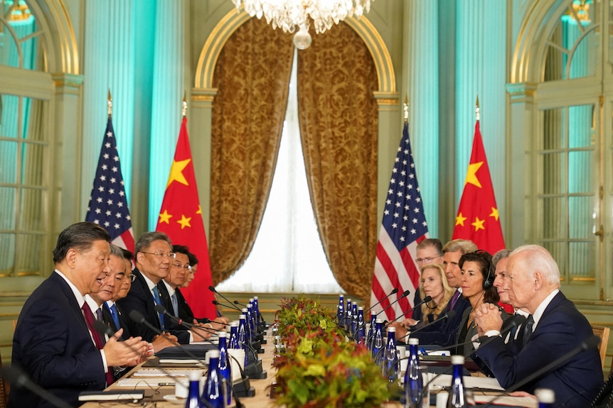 Biden and Xi sit opposite each other. Officials sit next to them at a long table. China and US flags hang next to a window.