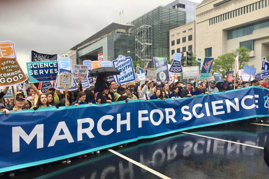 Scientists on a wet road in Washington DC carrying banners, in front a large banner as wide as the road saying March for Science