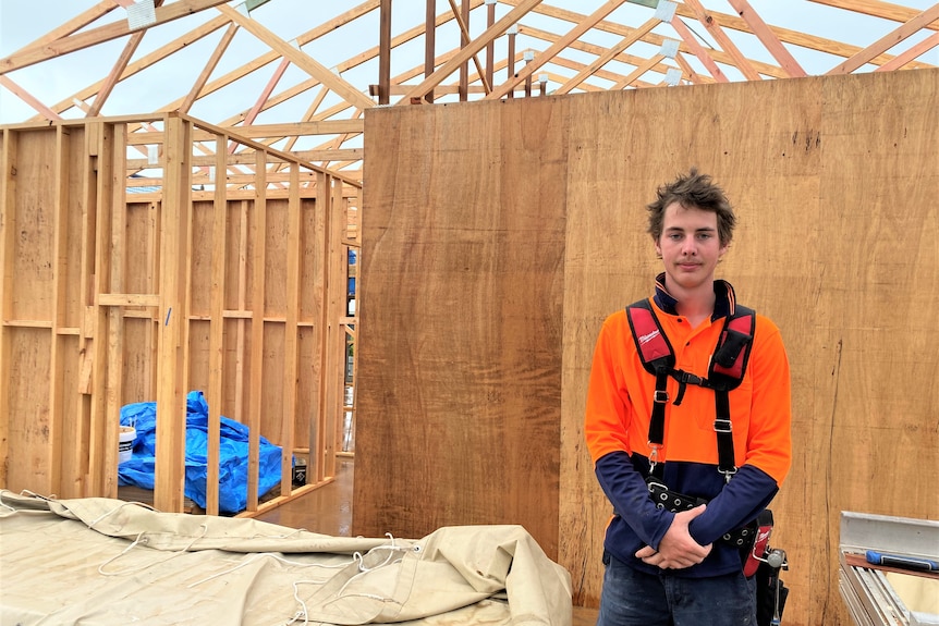 A young man standing on a building site wearing a tool belt and hi-vis orange shirt