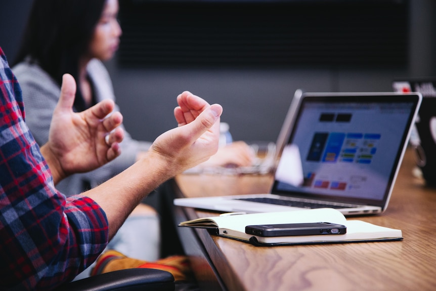 a laptop on the table, two work colleagues, one out of focus, the other only hands are visible