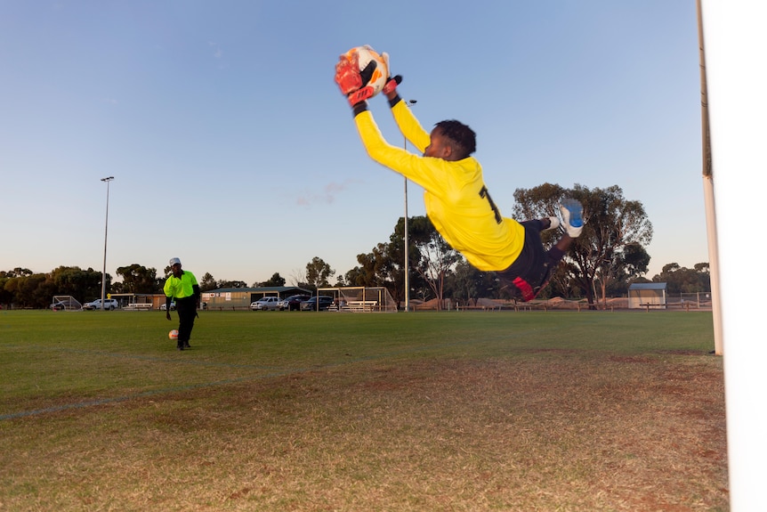 A young African Australian soccer player grabs a ball in mid air, while jumping across the pitch. The sky behind him is blue.