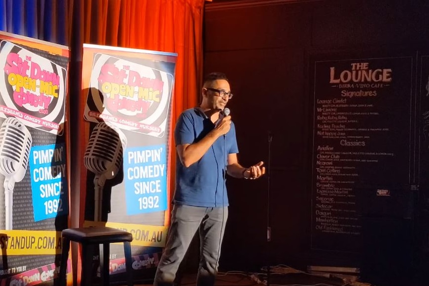 Vikesh stands on stage at a comedy club, with a microphone in hand.