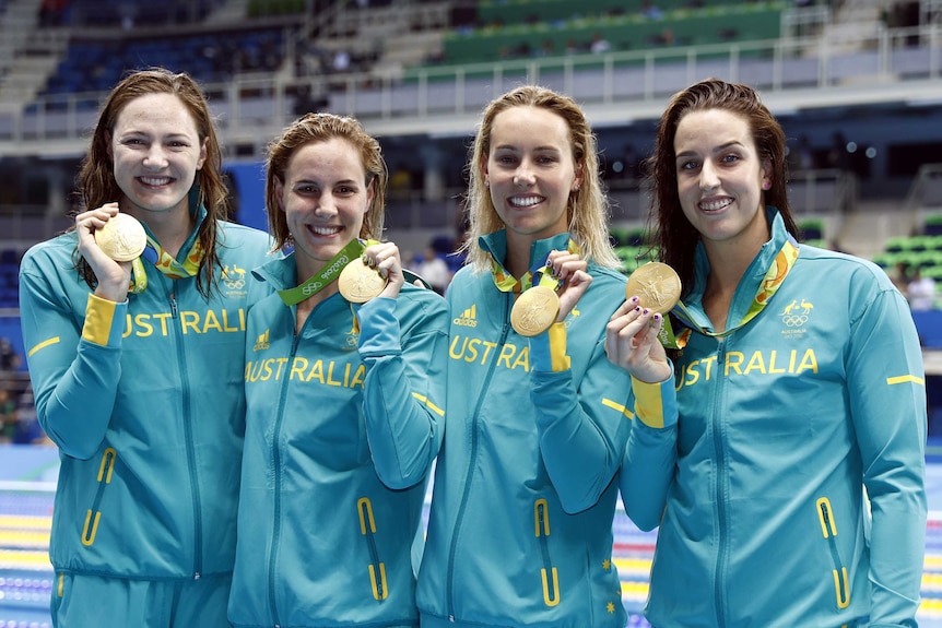 Four Australian swimmers stand holding their gold medals and smiling in the Olympic Aquatic Stadium.