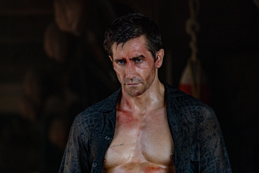 Jake looks down slightly with a serious expression, his shirt open over his bare chest and cuts and blood on his face.