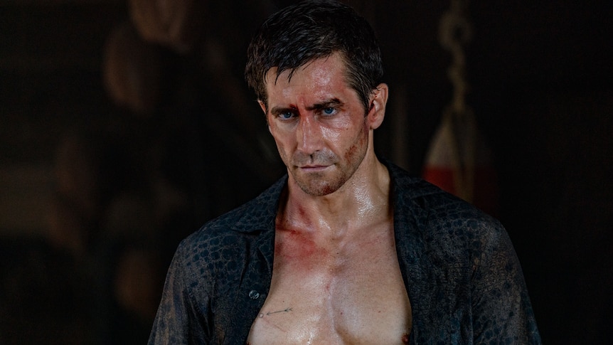Jake looks down slightly with a serious expression, his shirt open over his bare chest and cuts and blood on his face.