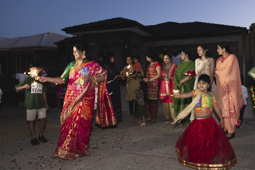 A woman wearing a bright saree and a child in a long skirt plays with sparklers as other women look on.