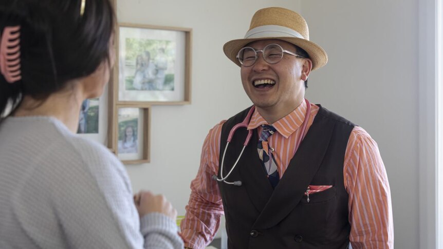 A man in a tan hat, glasses, pale orange pink shirt and waitcoast laughs. He has a stethoscope around his neck.