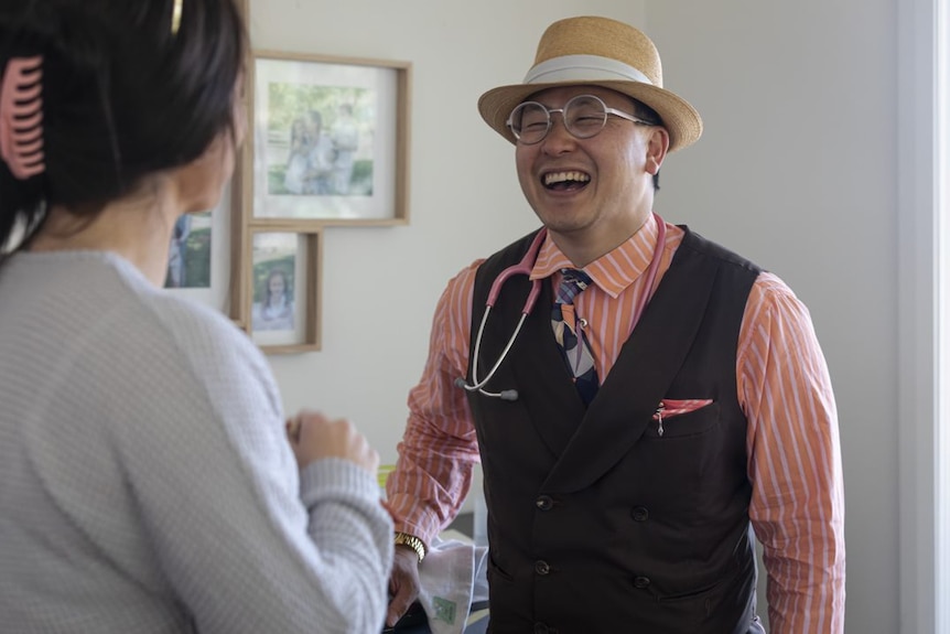 A man in a tan hat, glasses, pale orange pink shirt and waitcoast laughs. He has a stethoscope around his neck.