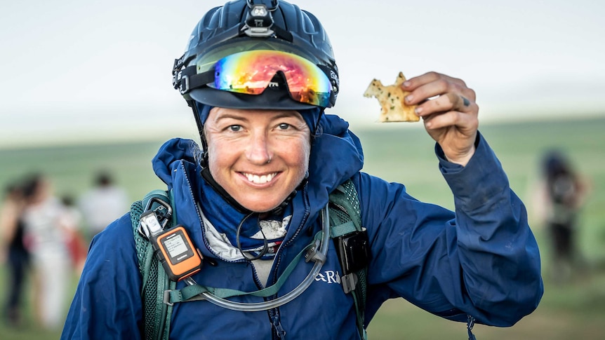 A woman out in a field wearing a horse riding helmet smiles holding a biscuit.