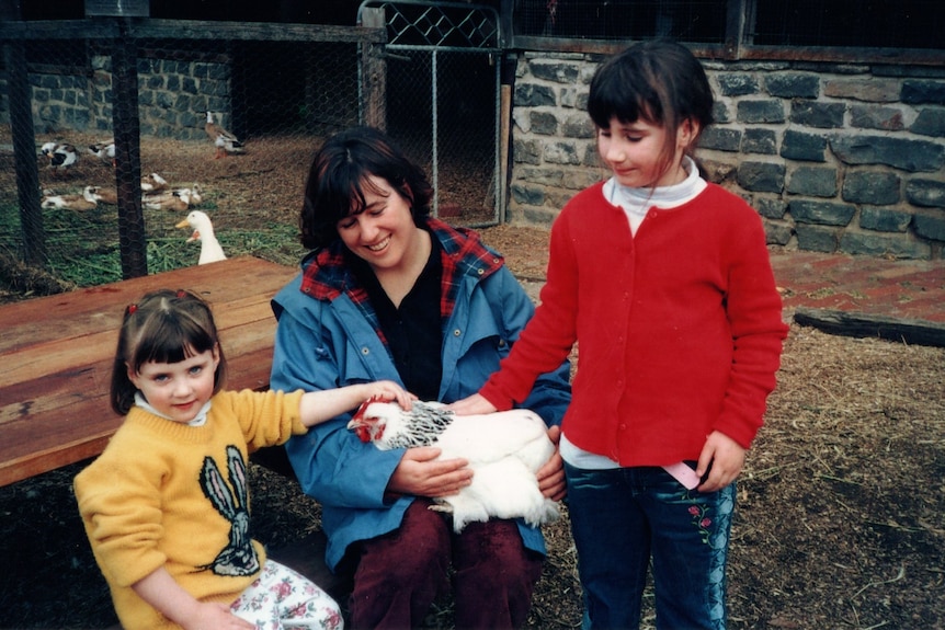 A woman holds a chicken on her lap while two girls pat the chicken.