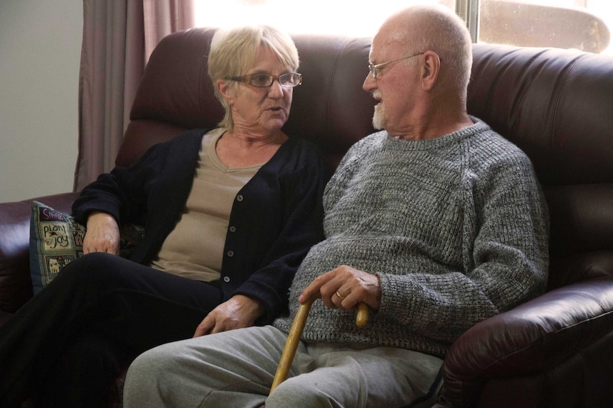 Elderly couple Christine and John Berry sit on a couch indoors talking to one another.