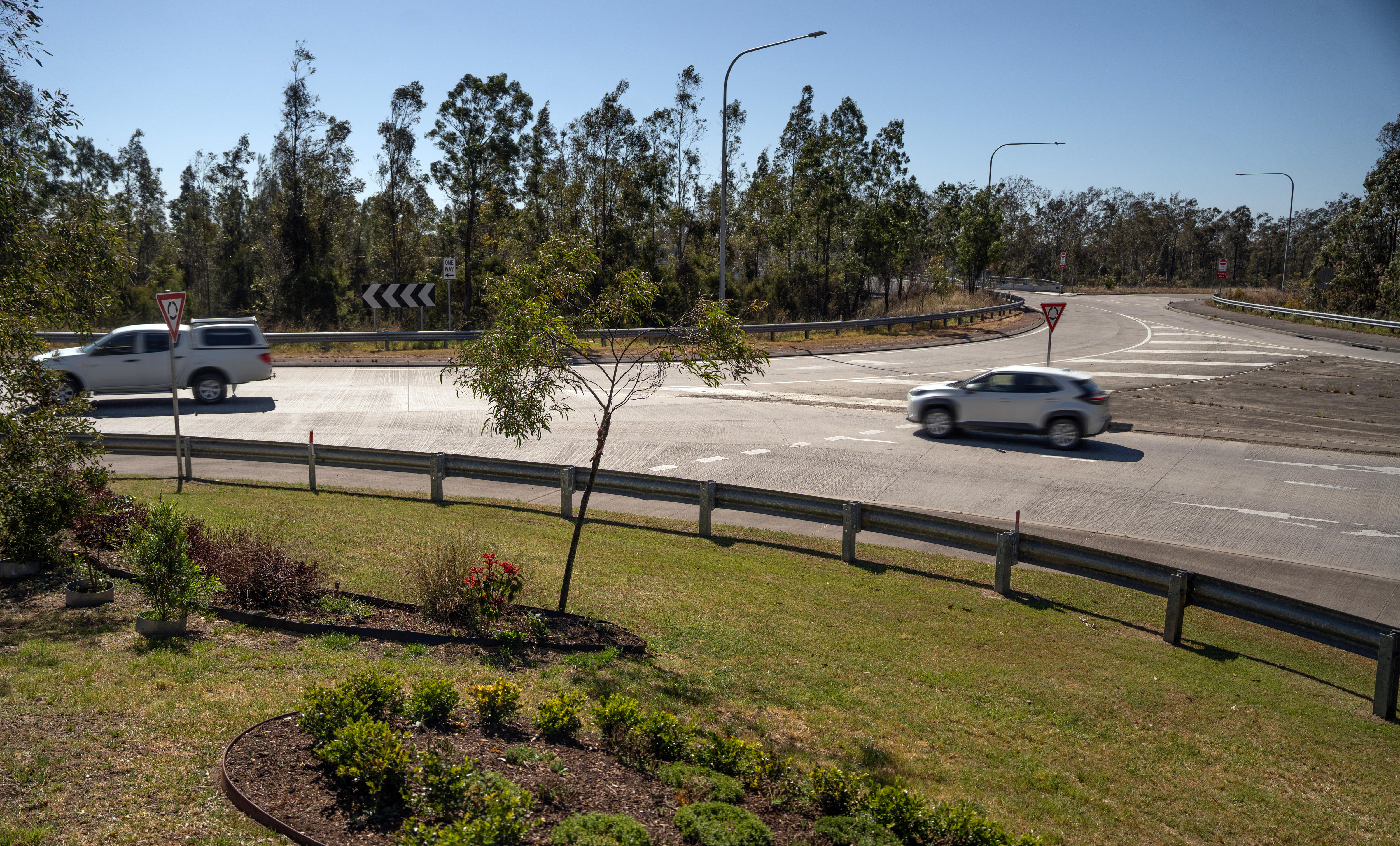 Two cars drive through a roundabout during the day. The road is surrounded by grass and trees in various points.