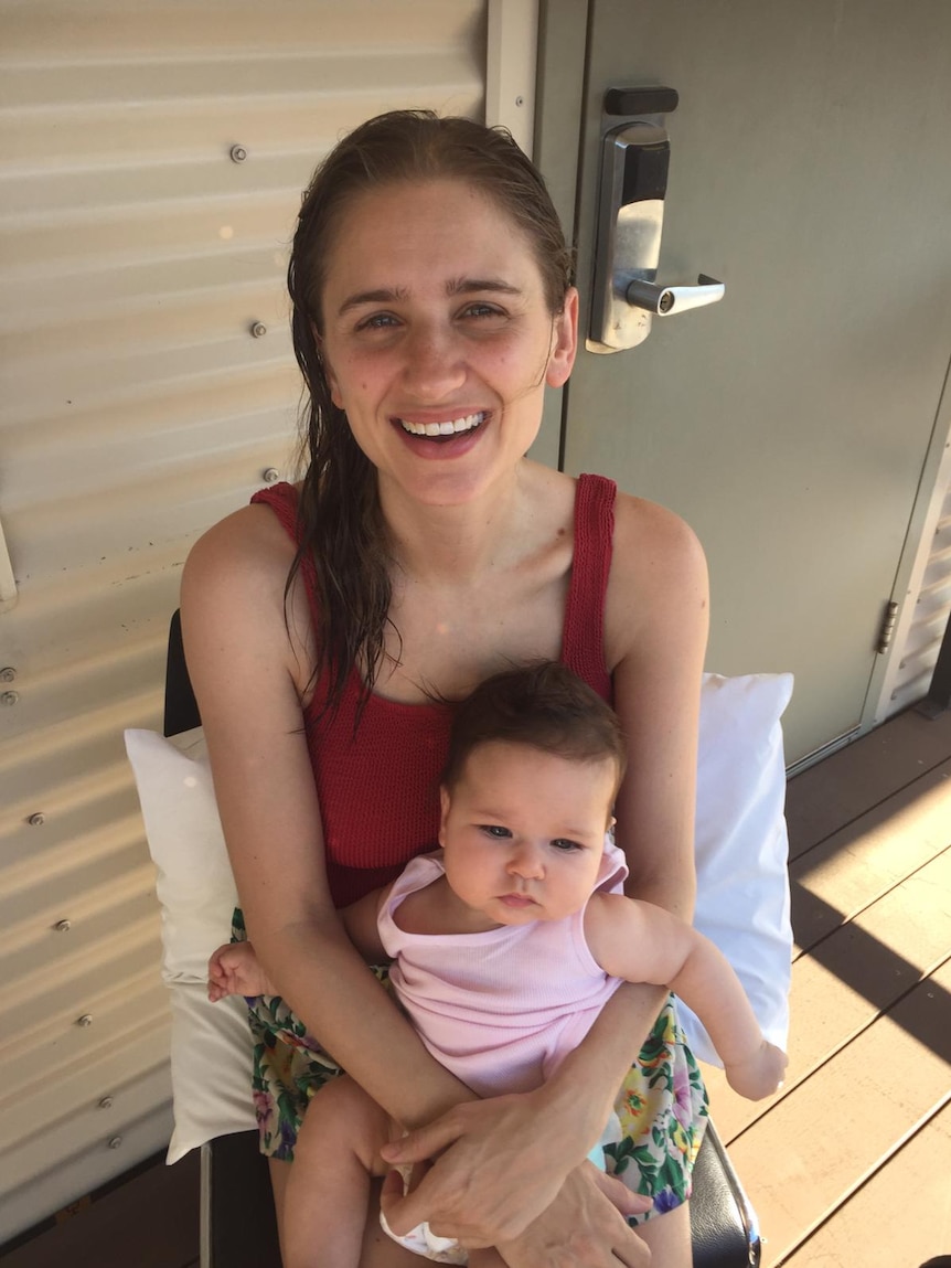 A photo of a young mother holding her baby daughter and smiling at the camera.