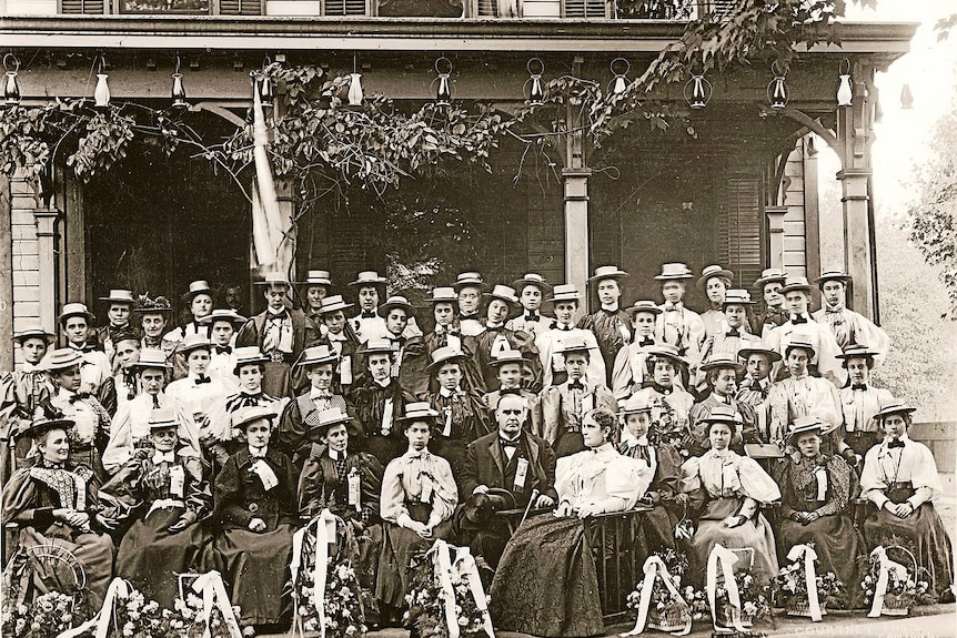 A black and white photo of a man sitting outside a house surrounded by women