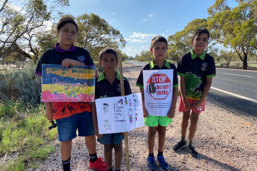 Four Indigenous children standing beside a road hold up a sign.