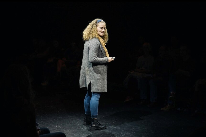 Lily Jones stands on a stage lit up with others standing in darkness behind her.