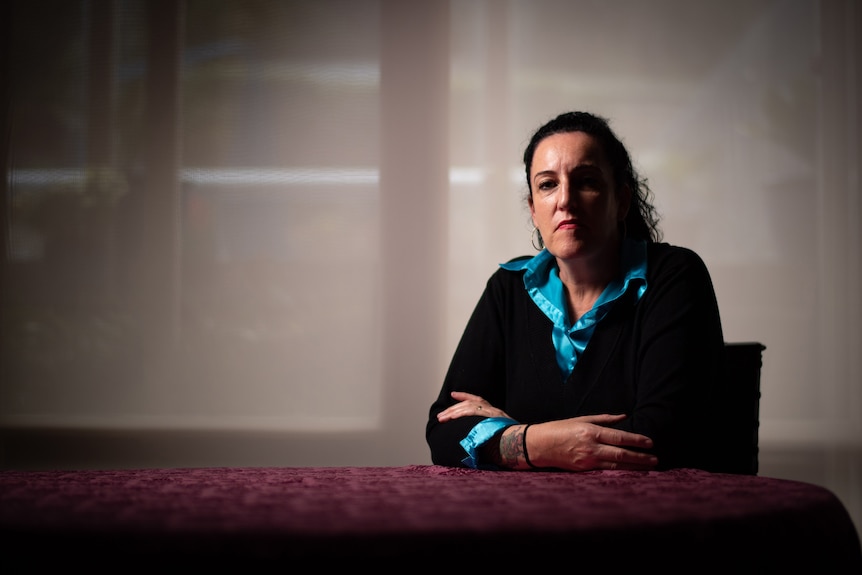 A woman sits a table, her arms folded. She has a neutral expression on her face, which is half in shadow.