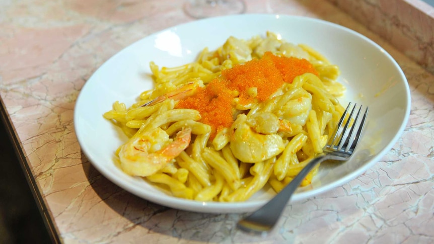 A seafood pasta dish with salmon roe and prawns, sitting on a restaurant bench with orange drink.