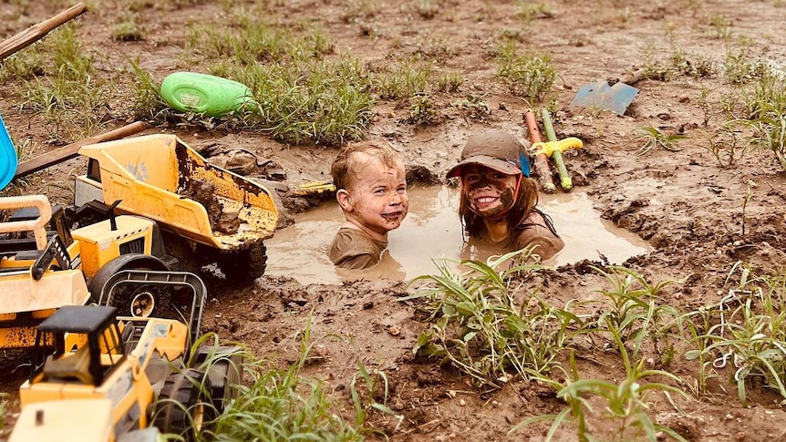 Two children are sitting in a deep hole filled with muddy water. They are smiling and have mud on their faces.